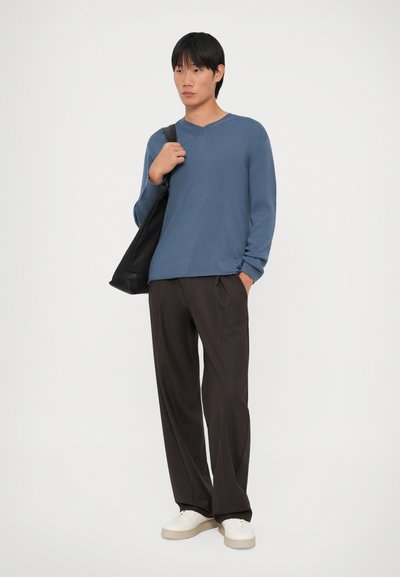 Μπλε ριγέ πουλόβερ με μακριά μανίκια και V λαιμόκοψη, σε συνδυασμό με χαλαρούς καφέ παντελόνες και λευκά αθλητικά παπούτσια. Το μοντέλο κρατά μια μαύρη τσάντα tote.