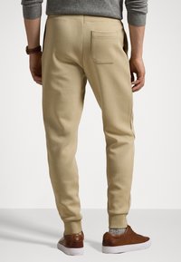 Mężczyzna ubrany w beżowe spodnie dresowe i brązowe skórzane trampki, stojący z rękami swobodnie opuszczonymi wzdłuż ciała, widoczny od tyłu.