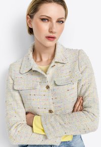 Struktureret, pastel tweedjakke med to forlommer, guldknaplukning og kravedesign. Båret over en gul top, stylet med korslagte arme.