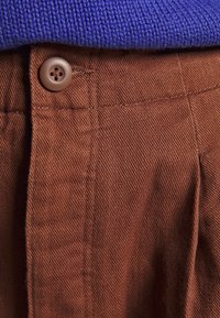 Bruna bomullbyxor med en strukturerad yta och knäppning, delvis täckta av en blå stickad tröja vid midjan.