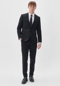 Black tailored suit with a smooth texture, featuring a single-button jacket, white dress shirt, and dark tie. Worn with black shoes.