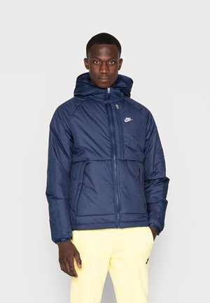 Marineblauwe puffer jack met een rits aan de voorkant, een capuchon en twee voorzakken. Kenmerkt zich door een textuurstof en een klein wit logo op de borst.