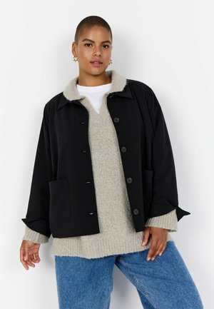 Person mit kurzen Haaren, der goldene Stern-Ohrringe trägt, schwarze, geknöpfte Jacke über grauem Pullover und blauer Jeans, steht vor weißem Hintergrund.