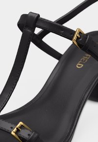 Μαύρο δερμάτινο σανδάλι με λεπτά λουριά και χρυσά μεταλλικά αγκράφες, κοντινή λήψη που δείχνει το λείο πέλμα και το όνομα της μάρκας σε χρυσά γράμματα.