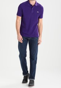 Camisa polo morada de manga corta y cuello, con un pequeño logo. Combinada con pantalones vaqueros azules y zapatos grises. Diseño y ajuste sencillo.