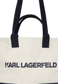 Kreminės ir tamsiai mėlynos spalvų drobės tote krepšys, turintis dvi rankenas ir ryškiai tamsiai mėlynai atspausdintą "KARL LAGERFELD" logotipą.