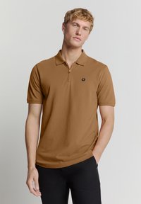 Bruine polo met een ritssluiting bij de kraag en korte mouwen. Heeft een zwart hexagonaal logo op de linkerkant van de borst. Gemaakt van glad, lichtgewicht stof.