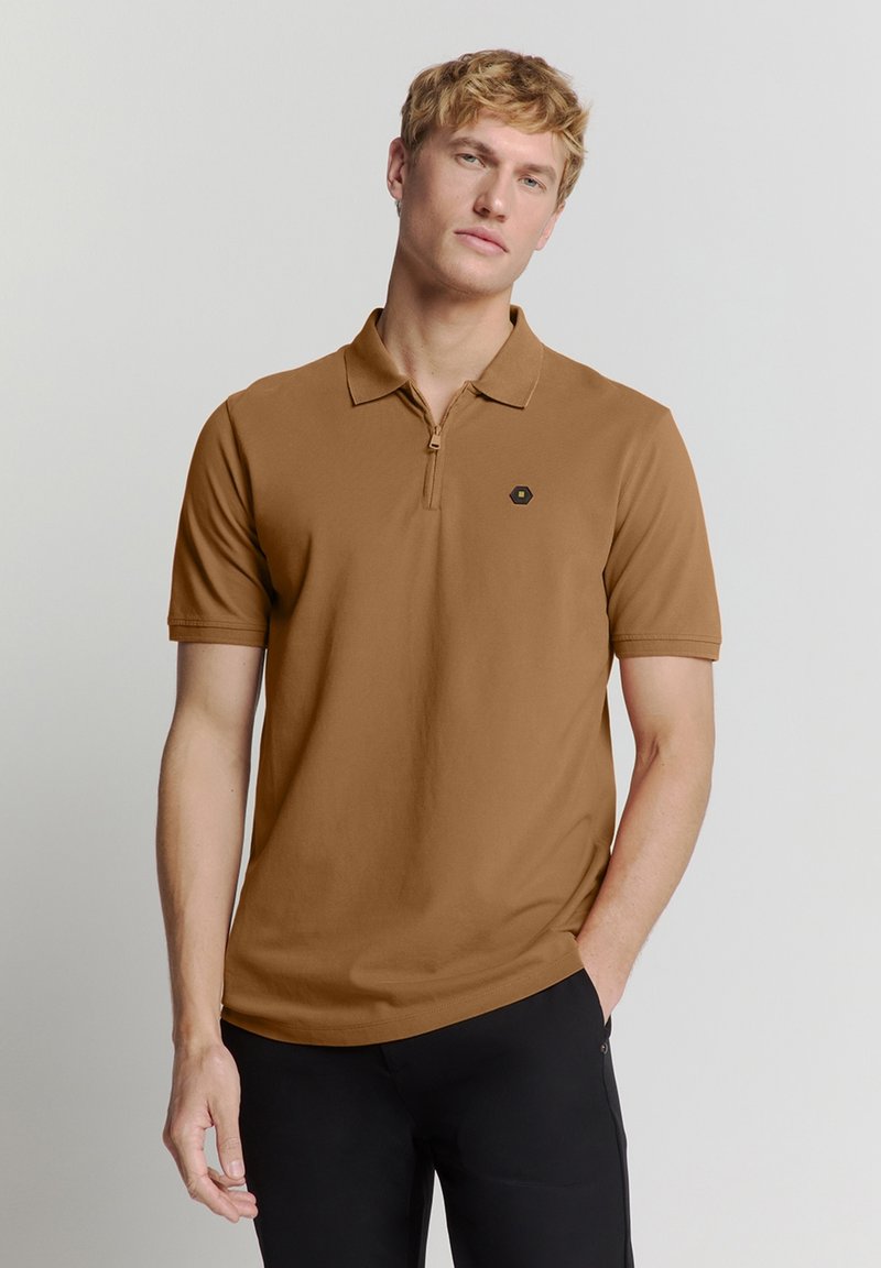 Bruine polo met een ritssluiting bij de kraag en korte mouwen. Heeft een zwart hexagonaal logo op de linkerkant van de borst. Gemaakt van glad, lichtgewicht stof.