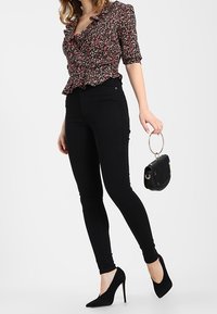 Mujer vistiendo pantalones ajustados negros de cintura alta, blusa floral con volantes, tacones negros altos, sosteniendo un pequeño bolso negro con un asa dorada redonda.