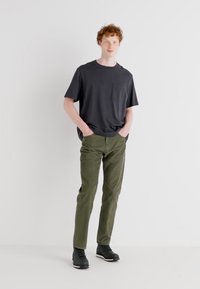 Camiseta de manga corta gris oscuro con un bolsillo en el pecho, combinada con pantalones verdes olivo y zapatillas negras, de pie sobre un fondo sencillo.