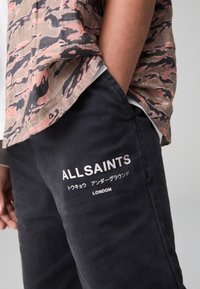 Čierne manšestrové nohavice s bielym logom "ALLSAINTS" a japonským textom na boku, skombinované so vzorovanou košeľou s krátkym rukávom v ružovej a čiernej farbe.