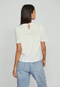 Witte kanten top met korte mouwen en een diamanten patroon, voorzien van een sleutelgatdetail op de rug, gecombineerd met lichtblauwe denim jeans.