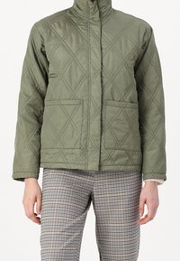 Quiltad olivgrön jacka med hög krage, diagonala sömmar och två framfickor; kombinerad med rutiga byxor. Texturerad tyg.