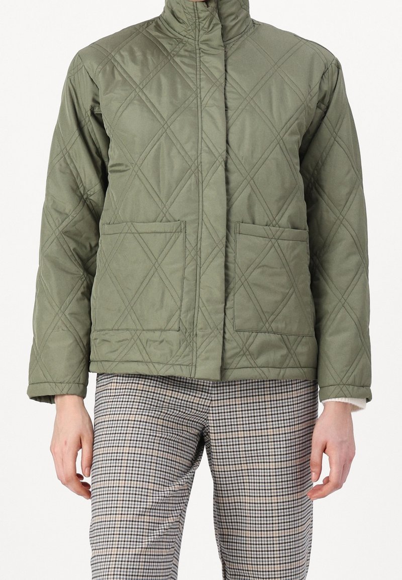 Quiltad olivgrön jacka med hög krage, diagonala sömmar och två framfickor; kombinerad med rutiga byxor. Texturerad tyg.