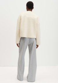 Giacca in pelliccia sintetica dalla texture cremosa con spalle scese, abbinata a pantaloni a gamba larga grigio chiaro a vita alta, vista da dietro.