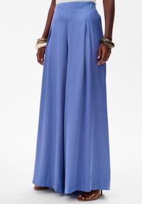 Pantalons à large jambe en tissu bleu clair avec une texture lisse, présentant des plis avant et une taille haute. Associé à des sandales à glisser marron.