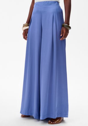 Lichte blauwe wijde broek van stof met een gladde textuur, voorzien van voorplooien en een hoge tailleband. Gecombineerd met bruine slidesandalen.