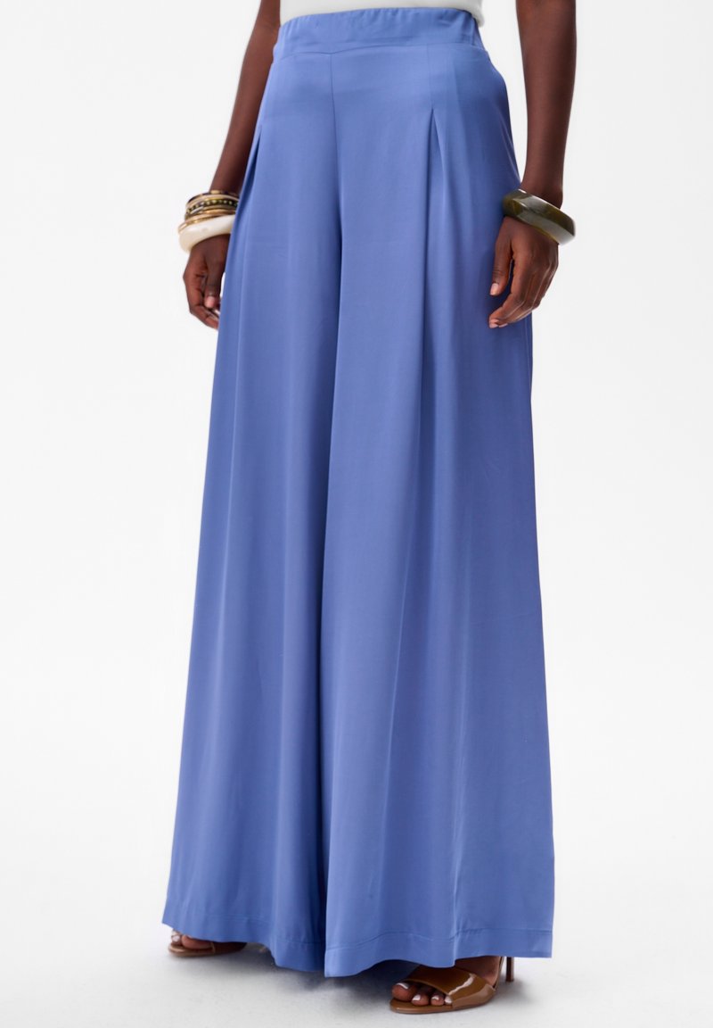 Pantalons à large jambe en tissu bleu clair avec une texture lisse, présentant des plis avant et une taille haute. Associé à des sandales à glisser marron.