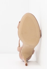 Beige high-heeled shoe leaning against a white wall, sole facing the camera, showing thin stiletto heel and ankle strap.