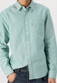 Camicia azzurra chiara a maniche lunghe con bottoni, realizzata in un tessuto morbido, presenta una tasca sul petto e colletto classico, con bottoni bianchi.