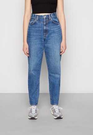 Person trägt hoch taillierte blaue Jeans, ein schwarzes kurzes Oberteil und silberne Sneaker und steht vor einem schlichten hellen Hintergrund.