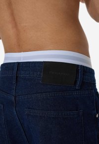 Προβάλλονται σκούρα μπλε τζιν παντελόνια, με λευκή και γκρι ριγωτή μέση και μαύρη δερμάτινη ετικέτα στην πίσω τσέπη.