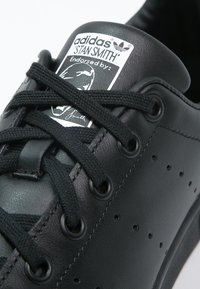 Svart lädersneaker från Adidas Stan Smith med perforeringar, svarta skosnören och en etikett på tungan med en logotyp och godkännande grafik.