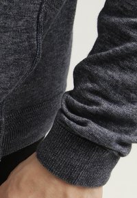 Nahaufnahme eines Handgelenks, das einen dunkelgrauen gestrickten Pullover mit einem gerippten Bund und strukturiertem Stoff trägt.