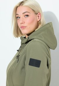Olivově zelená bunda s kapucí s hladkým povrchem, nastavitelnými šňůrkami a černou nášivkou s logem na rukávu. Odráží moderní, minimalistický design.