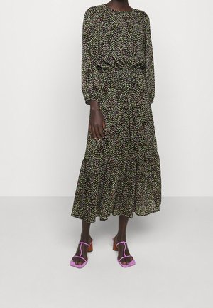 Kvinde iført en sort blomsterprintet midikjole med lange ærmer og en markeret talje samt lyserøde hælede sandaler med åben tå, stående på et hvidt gulv.