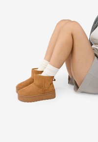 Botas de ante marrón hasta el tobillo con una suela de plataforma gruesa y una costura lateral. Llevadas con calcetines blancos acanalados, piernas cruzadas, sobre un fondo blanco liso.