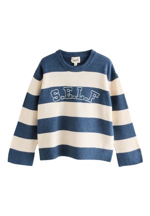 Giovane donna dai capelli ricci che indossa un maglione a righe blu e bianche con le lettere "S.E.L.F", appoggiata a un veicolo verde chiaro all'aperto.