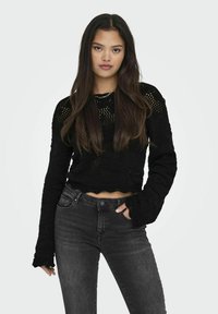 Maglione nero corto con un intricato pattern di maglia e maniche lunghe, abbinato a jeans scuri a vita alta. Il tessuto texture crea un effetto a strati.