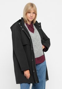 Giacca nera impermeabile con cappuccio, dotata di una zip frontale, due tasche e una vestibilità comoda, indossata sopra un maglione grigio a maglia e una camicia bordeaux.