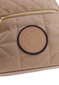 Bolso de nylon acolchado en color beige que presenta un cierre dorado, un parche de logo en cuero texturizado color marrón claro con un contorno azul oscuro, y costuras en contraste.