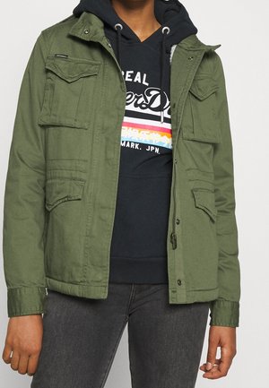 Henkilö pukeutunut oliivinvihreään takkiin mustan hupullisen paidan päällä, jossa valkoista ja värikästä graafista tekstiä, yhdistettynä tummanharmaisiin farkkuihin.