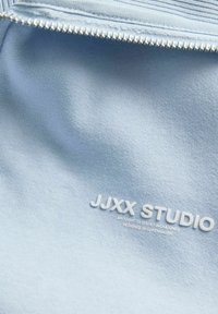 Svetlo modra tkanina z mehko teksturo, z detajli z zadrgo in embosiranim logotipom "JJXX STUDIO" v belem, nameščenim na materialu.