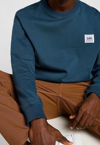 Sudadera de cuello redondo en azul marino, hecha de algodón, con puños acanalados y un pequeño parche de logo. Combinada con pantalones marrones texturizados.