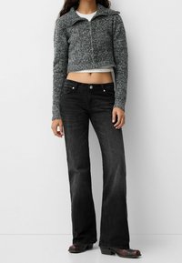 Cárdigan corto de punto gris con un gran cuello, combinado con vaqueros de pierna ancha negros y botas de tobillo negras. Diseño sencillo, sin patrones visibles.
