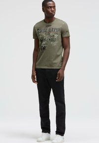 Olive green T-shirt with graphic print, crew neck, short sleeves, paired with black pants and white sneakers. Smooth texture, relaxed fit.