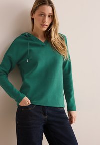 Grön huvtröja med v-ringad hals, strukturerad tyg, långa ärmar och dragsko. Matchas med mörkblå jeans med gul sömnad.