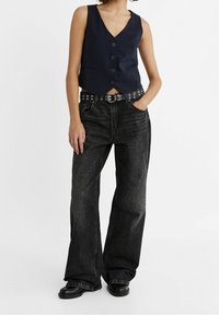 Marineblaue ärmellose Weste mit V-Ausschnitt und Knopfverschluss, kombiniert mit hochgeschnittenen, dunkelgrauen weit geschnittenen Jeans, akzentuiert durch einen Nieten Gürtel.