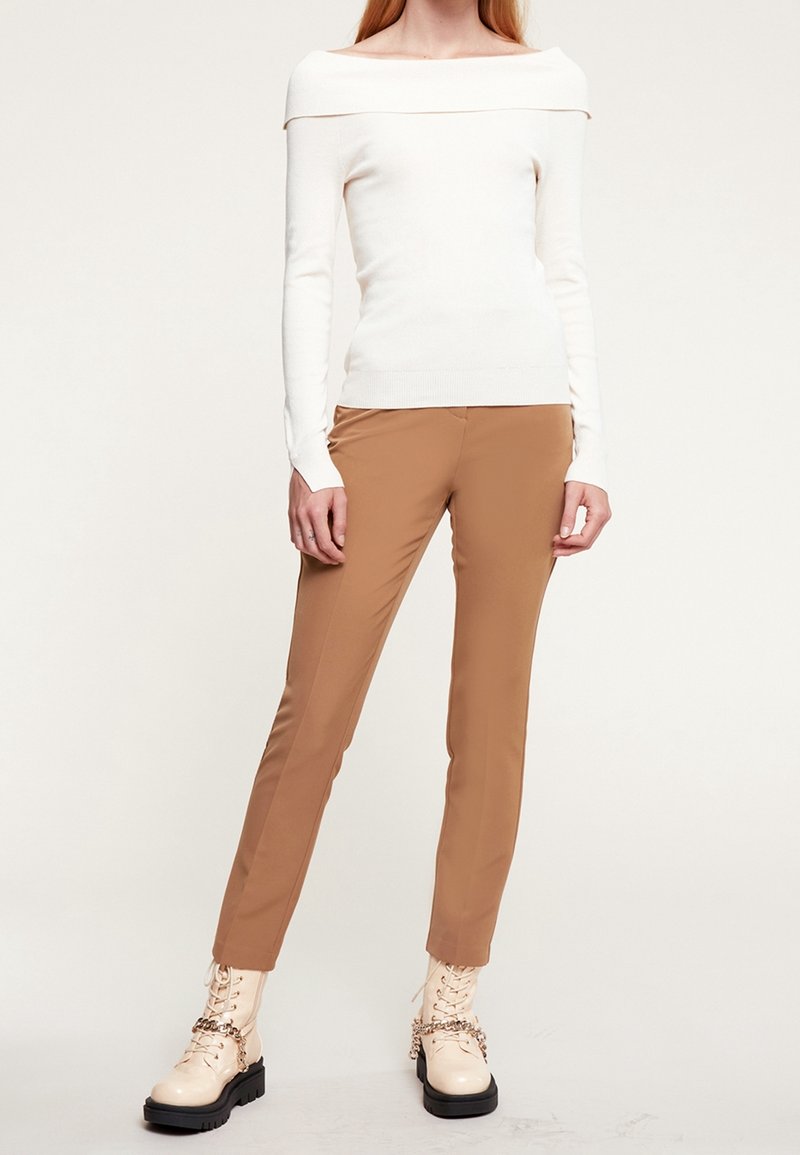 Pull blanc cassé à épaules dénudées, silhouette ajustée, associé à un pantalon camel sur mesure et des bottes de combat crème épaisses avec lacets et détail chaîne.