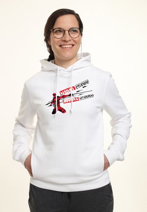 Uśmiechnięta kobieta w okularach ubrana w białą bluzę z kapturem z czerwono-czarną grafiką Marvel Studios, z rękami w kieszeniach na jednolitym tle.