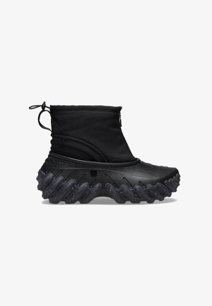 Bottes noires à la cheville avec une tige en textile doux et une semelle en caoutchouc texturée et ondulée. Équipées d'une fermeture éclair et d'un cordon réglable pour un ajustement parfait.