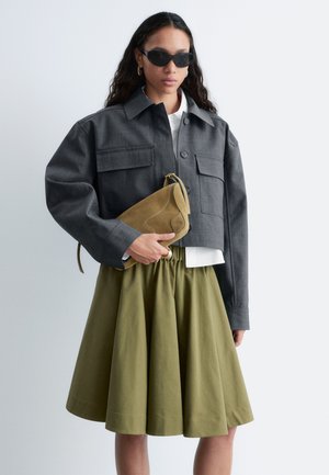 Kobieta w czarnych okularach przeciwsłonecznych, krótkiej ciemnoszarej kurtce, białej koszuli i oliwkowozielonej spódnicy, trzymająca beżową zamszową torebkę na jednolitym tle.