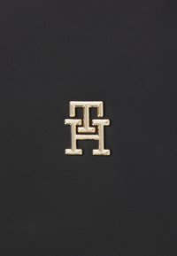 Texture in nylon nera con un logo dorato in rilievo prominente, caratterizzato da un design intrecciato "TH", evidenziando un'estetica elegante e moderna.