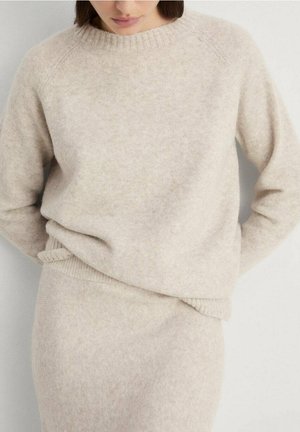 Femme portant un pull en laine beige doux et une jupe assortie, les mains derrière le dos, sur un fond clair uni.