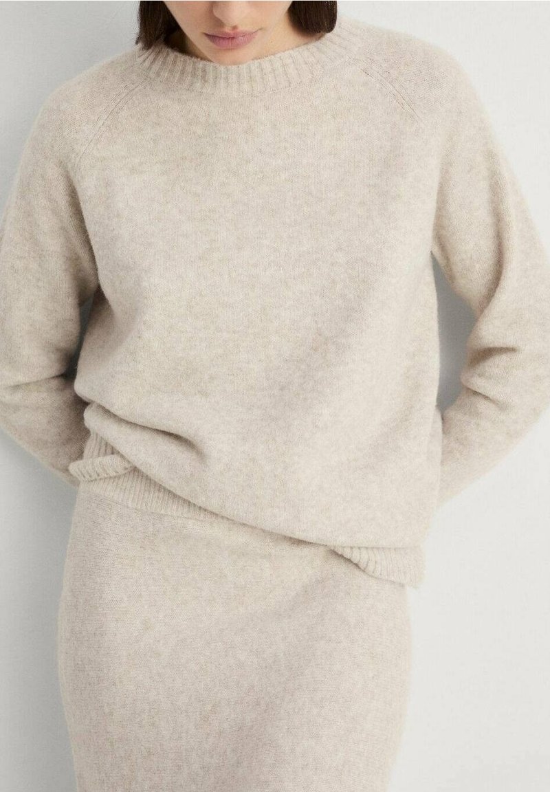 Femme portant un pull en laine beige doux et une jupe assortie, les mains derrière le dos, sur un fond clair uni.