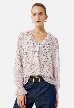 Femme portant un chemisier léger rose clair transparent avec des pois noirs, un devant froncé et des poignets, associé à un jean bleu foncé et une ceinture marron.